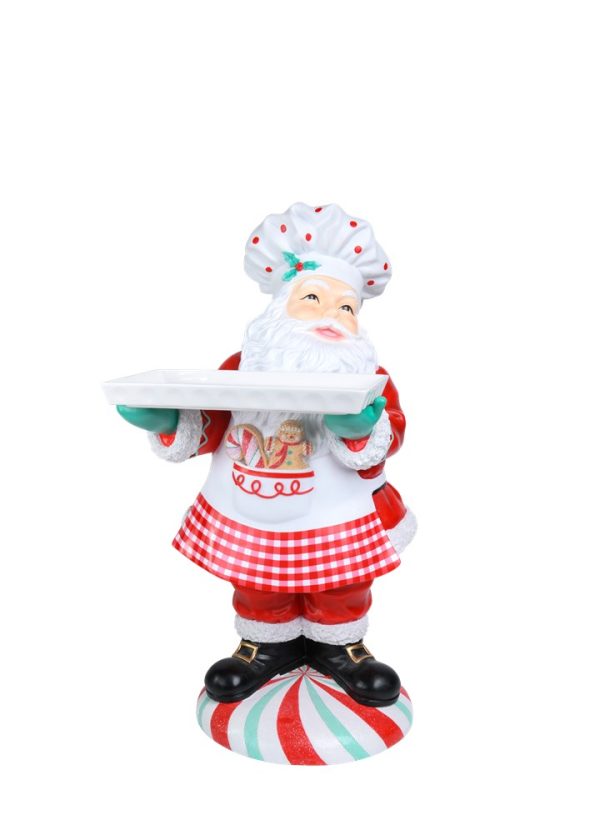 A festive Santa Claus figurine dressed as a chef, holding a white serving tray. The Santa figure wears a white chef's hat with red dots, a white apron with a red gingham skirt overlay, and black boots, standing on a red, white, and green striped candy cane-patterned base against a plain white background.
