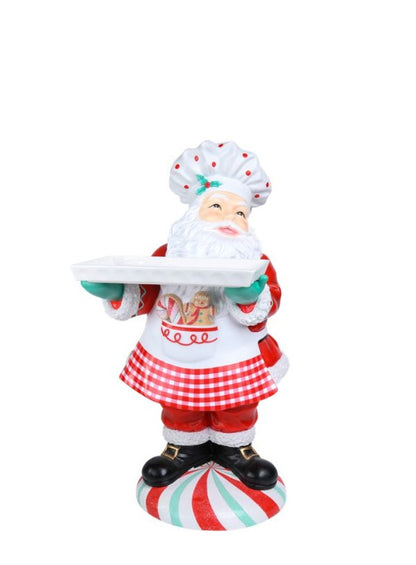 A festive Santa Claus figurine dressed as a chef, holding a white serving tray. The Santa figure wears a white chef's hat with red dots, a white apron with a red gingham skirt overlay, and black boots, standing on a red, white, and green striped candy cane-patterned base against a plain white background.