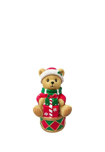 A small teddy bear Christmas ornament wearing a red and white Santa hat, a green and red striped sweater, and holding a red and green gift box, sitting on a matching drum base against a white background.