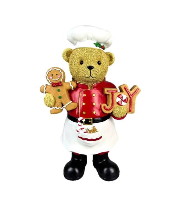 A festive teddy bear figurine dressed as a chef, wearing a white hat and apron with a red jacket, holding a gingerbread man and the letters 'J' and 'Y' to form 'JOY'.
