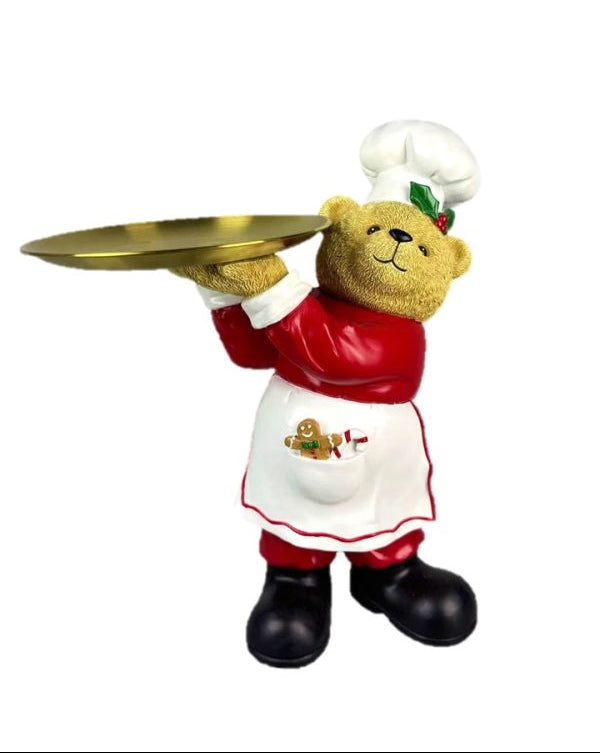 A teddy bear figurine dressed as a chef, holding a gold tray. The bear is wearing a white chef's hat, a red jacket, and a white apron with red trim, standing on black boots against a plain white background.
