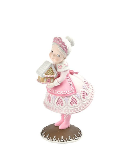A decorative figurine of Mrs Claus in a festive pink pastel dress, holding a gingerbread house, standing on a brown base against a white background.