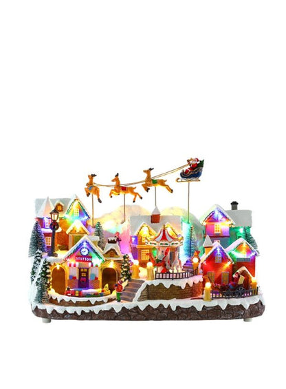 A festive miniature Christmas village scene with glowing houses, snow-covered roofs, reindeer on poles, Santa in a sleigh, and warm lights, shown on a plain white background.